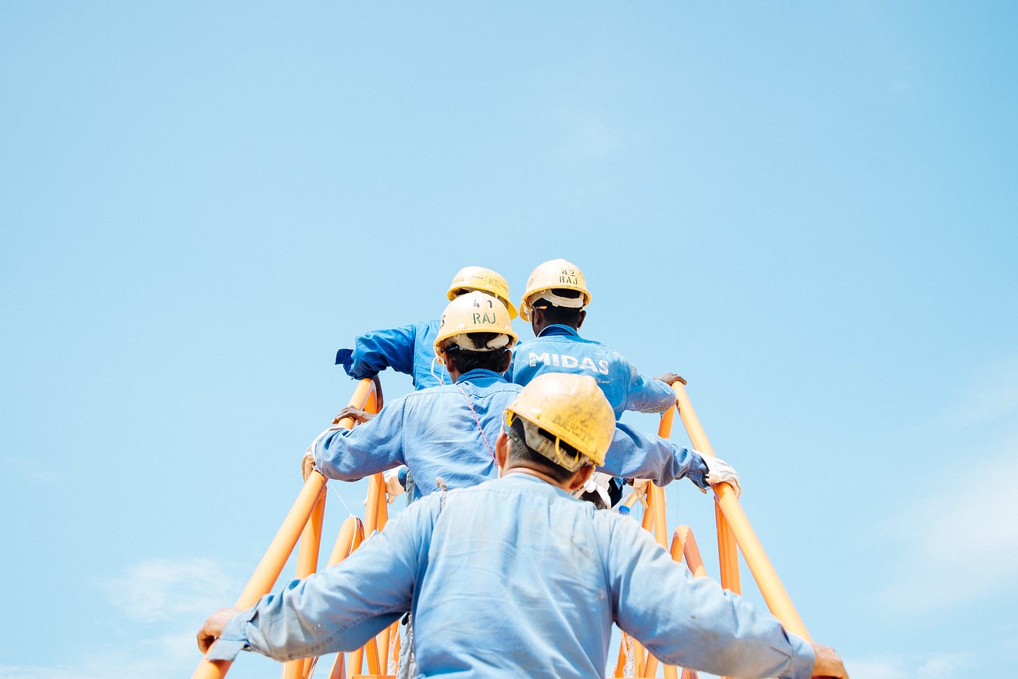 A photo of four workers, seen from behind as they climb a staircase with yellow metal railings towards a blue sky. The worker are wearing blue shirts and yellow hard hats.