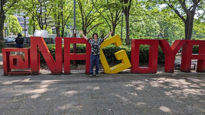 Photo of Rey smiling with arms spread wide in front of large metal letters spelling "Nagoya", the name of the city