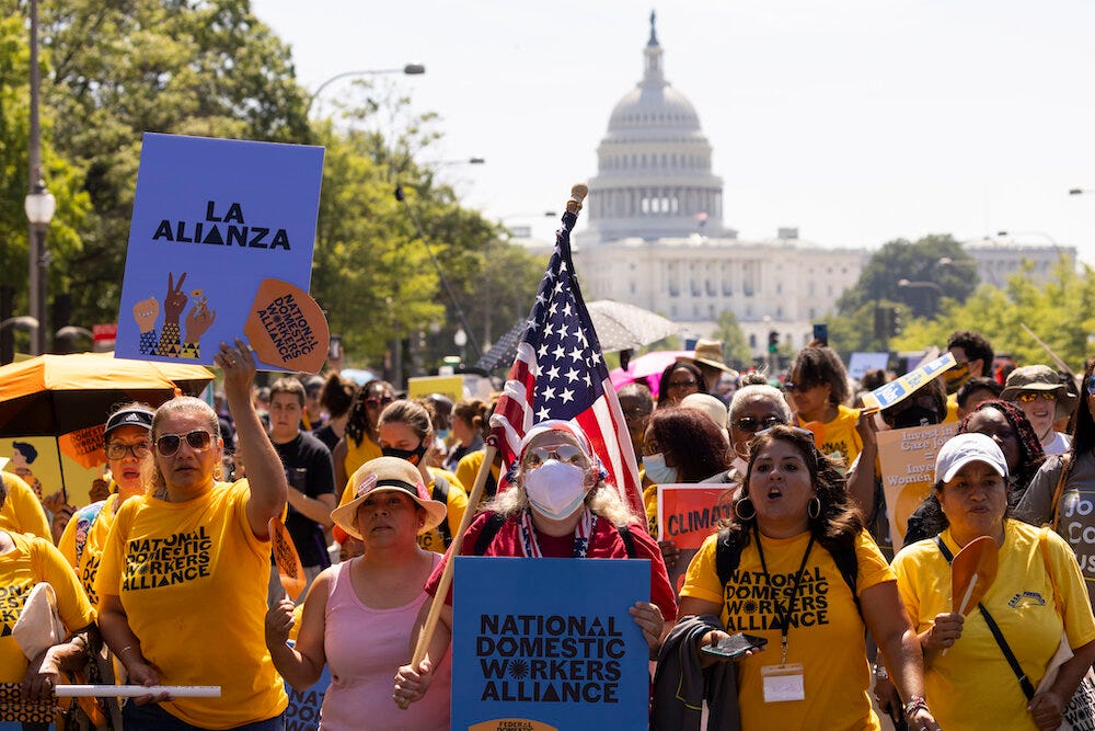 A photo of a people in a street, marching away from the U.S. Capitol in the background. Many wear yellow National Domestic Workers Alliance shirts. Someone holds an American flag. Another holds a sign that says “LA ALIANZA.”