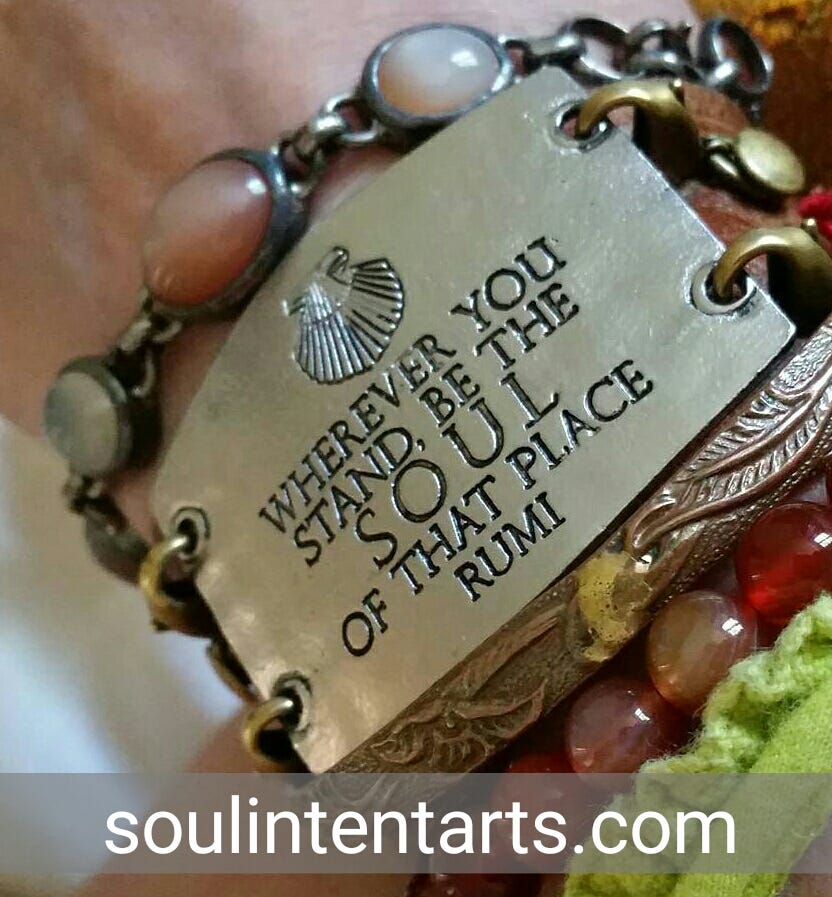A wide metal bracelet reading "Where you stand, be the soul of that place, Rumi" "soulintentarts.com"
