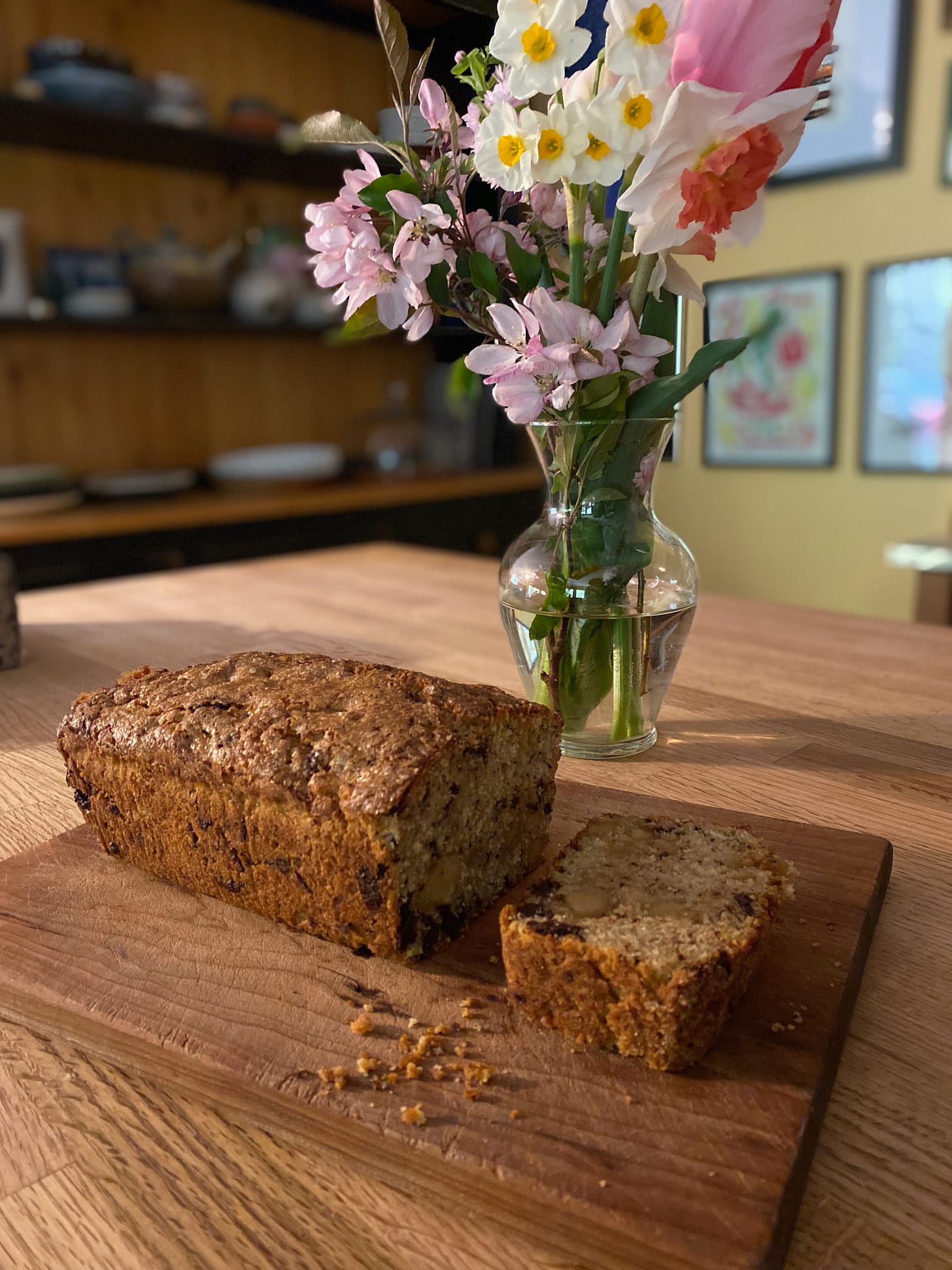 The scone loaf, with one slice cut off, sits on a wooden cutting board. A vase of spring tulips and daffodils sits behind it. The kitchen island is dappled with light.