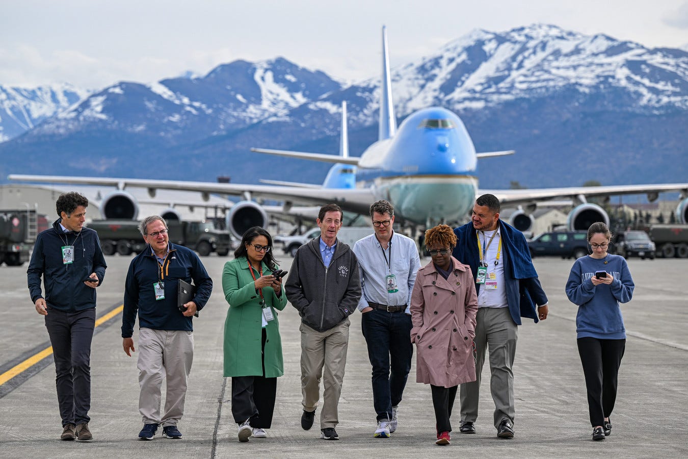 White House Deputy Chief of Staff Bruce Reed and White House Press Secretary Karine Jean-Pierre walking with reporters on a tarmac with Air Force One and mountains in the background.