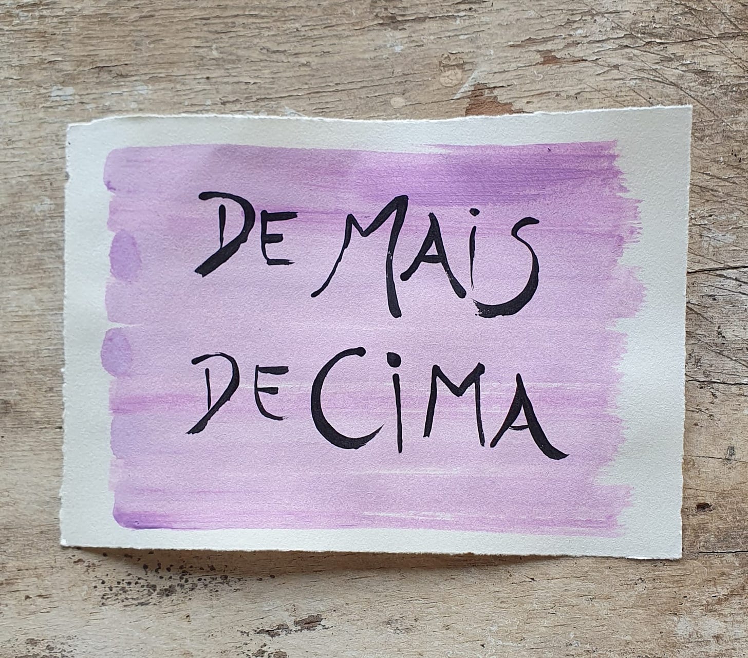 Fotografia de um papel pintado de lilás e o título De mais de cima pintado em preto em cima de uma base de madeira.