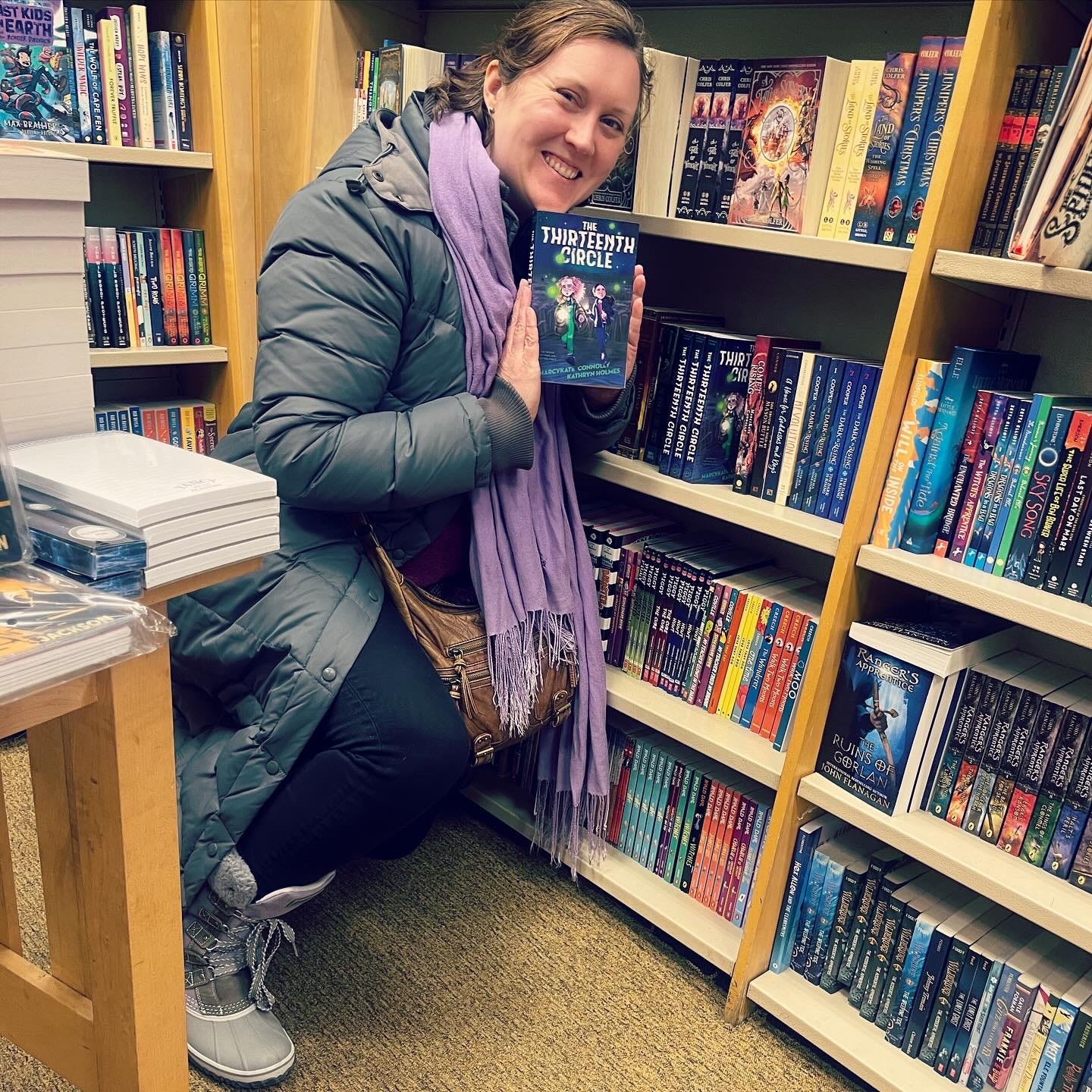 Kathryn poses with a paperback copy of The Thirteenth Circle, crouched next to the shelf in Barnes & Noble.