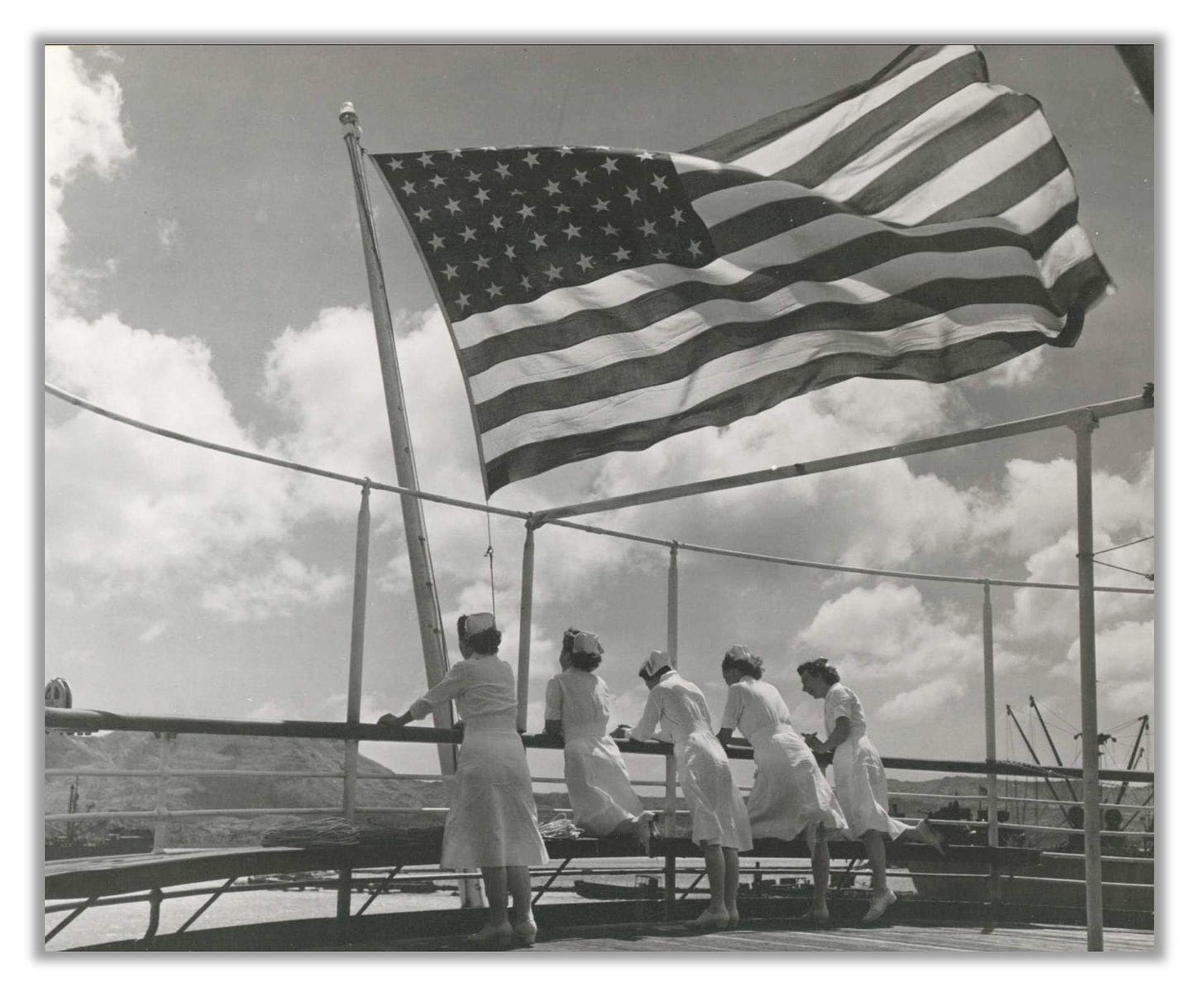 Navy Nurses aboard USS Solace (1945). A large United States flag flies above their heads as they lean over the rail.