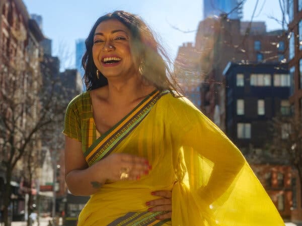 In a portrait, Tashnuva Anan Shishir is laughing while standing outside on a city street on a sunny day, with buildings in the background. She wears a yellow sari with green trim and has her left hand on her waist, with her other hand blurred as it moves in front of her.
