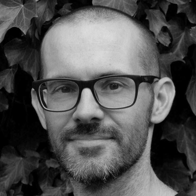 Black and white headshot of Rhodri, who is a white man with very short hair, dark-rimmed glasses, and a beard. He's standing in front of a wall of leaves, smiling slightly with his mouth closed.
