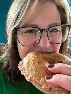 Me, a white woman with brown hair and a blonde fringe, is holding a bagel to her mouth and smiling. I'm wearing black rimmed glasses and have a green jumper. The bagel has salmon pate oozing out of it at the sides. I'm doing a mixture of biting and smiling.