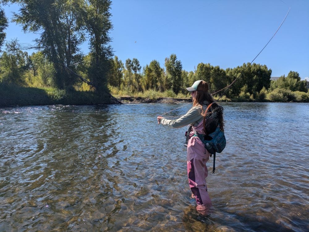 a young woman in pink waders, casting a fishing line in a river with trees along the bank