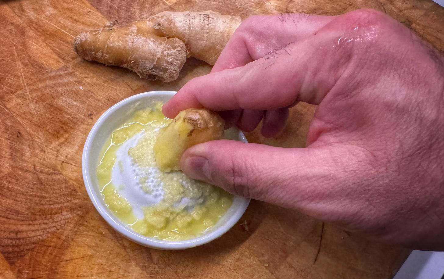 A hand grating a large piece of ginger on a ceramic ginger grater. Ground ginger and ginger juice pool around the edge of the grater.