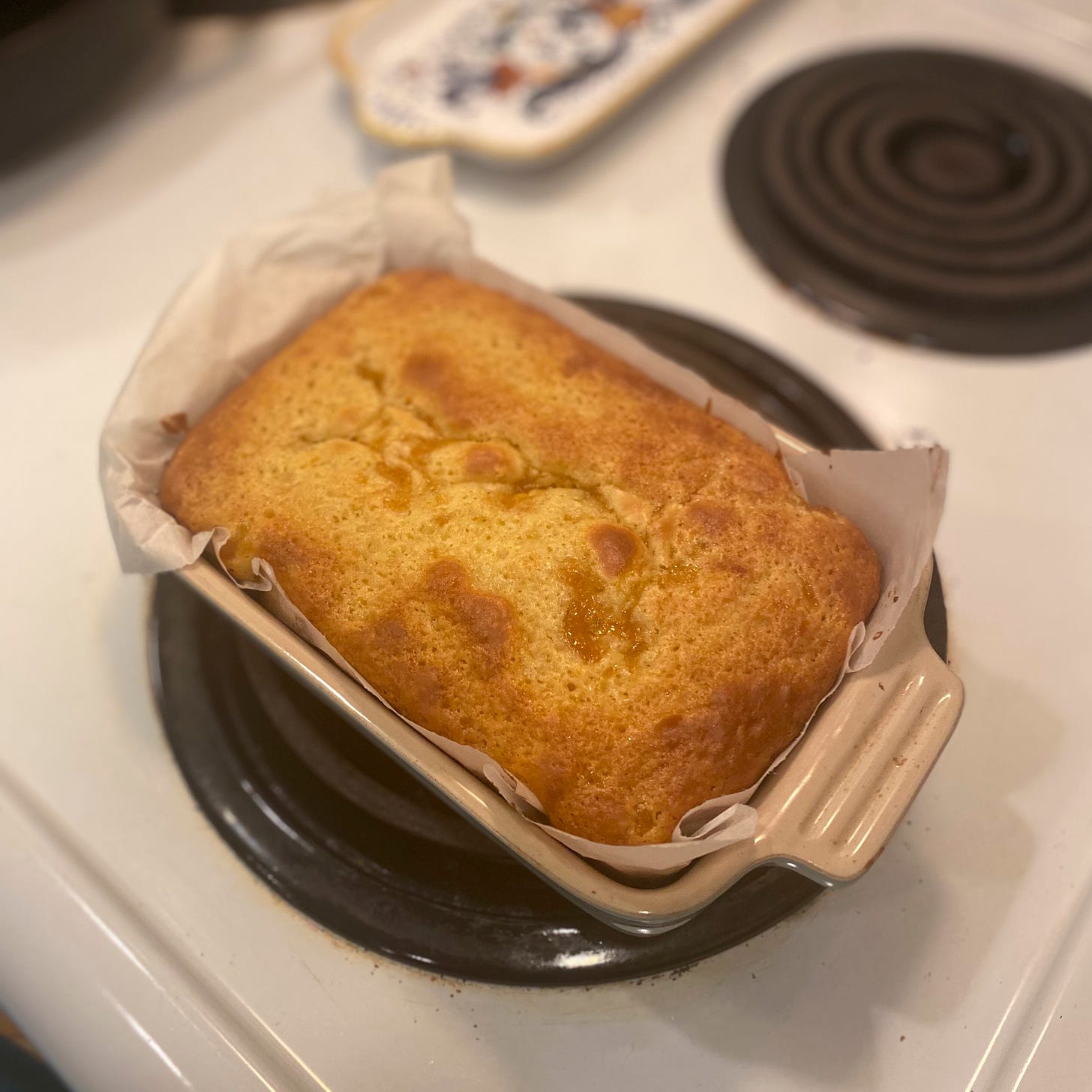 On the stovetop, a small Le Creuset loaf pan filled with parchment paper and a golden-brown cake, slightly sunk in the middle with peach jam.