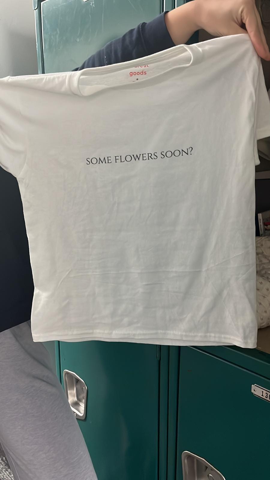 A hand holds up a small white t-shirt with the words "SOME FLOWERS SOON?" printed in black capitals across the middle