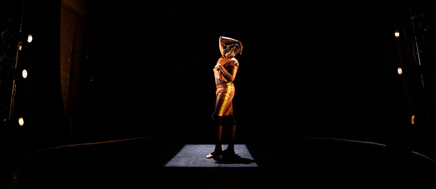 A Black disabled man is standing in a square light on a darkened stage. He is holding one hand over his face and head, the other close in to his body. He wears brown and yellow leather-looking garments.