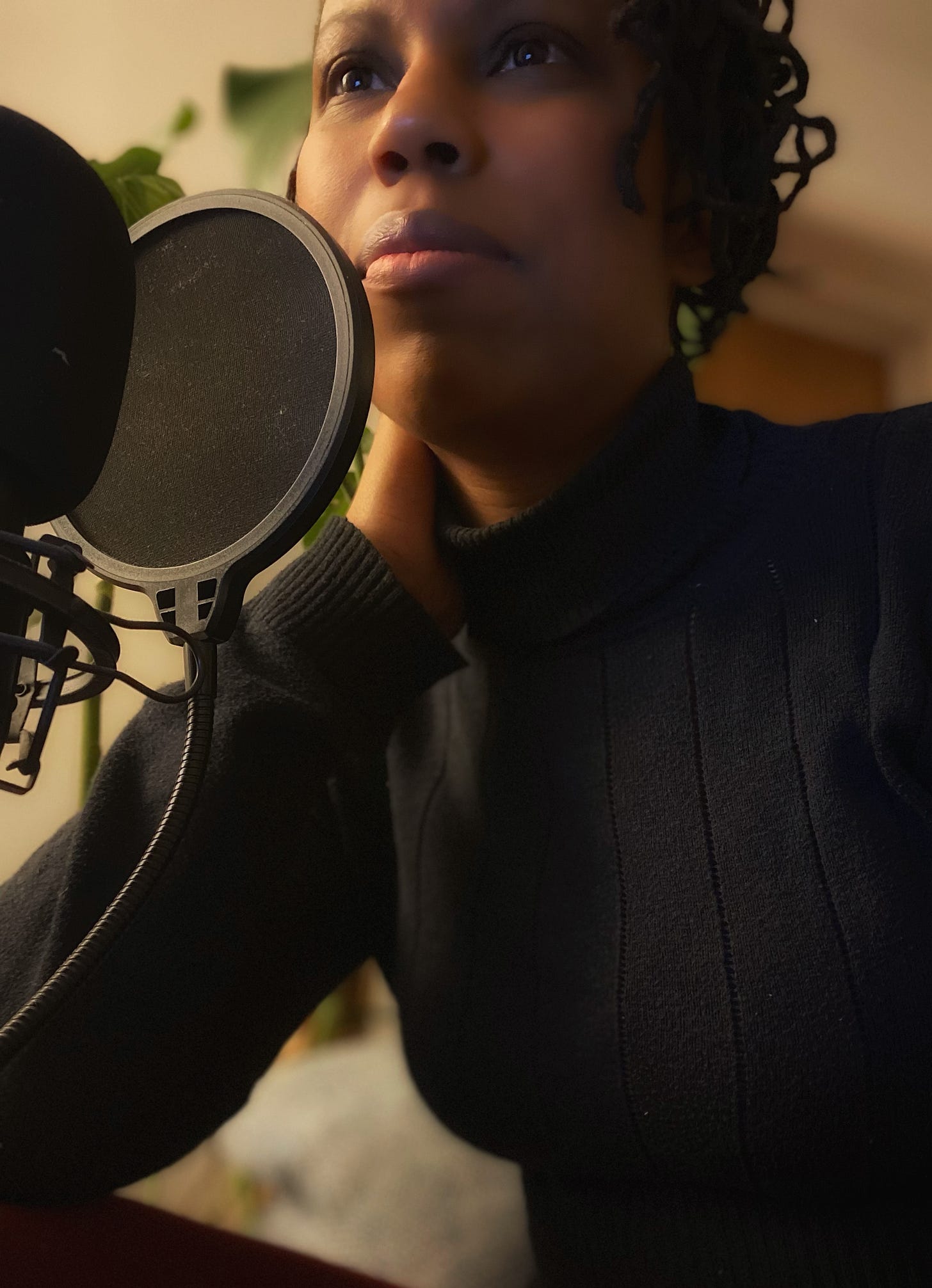Sandra is a Black femme with short black locs, wearing a black turtleneck, sitting in front of a podcast mic