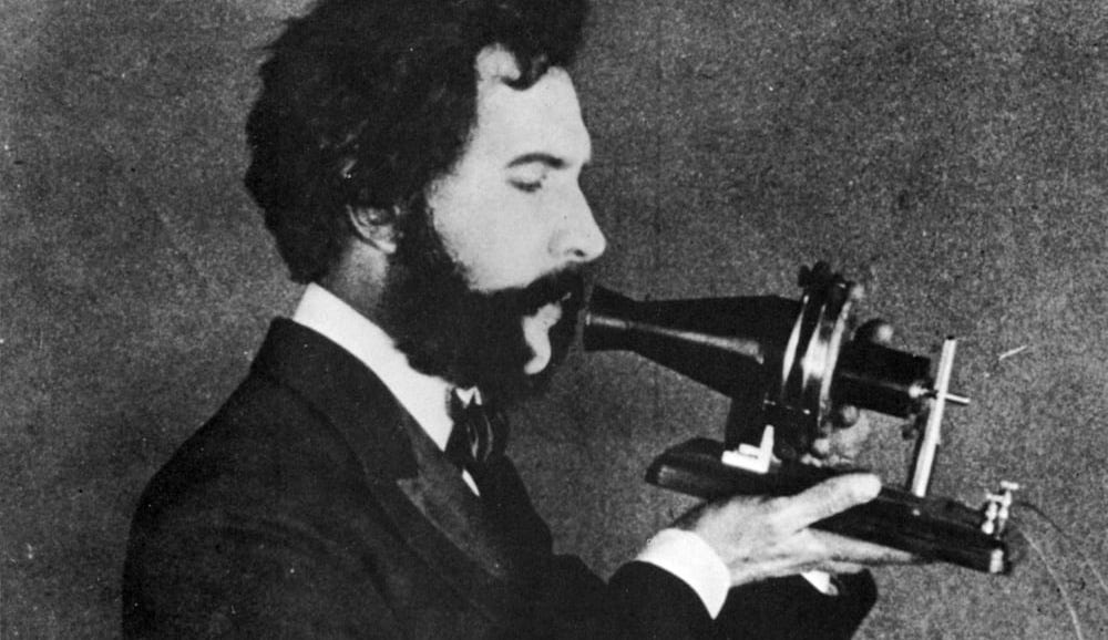 Imagem em preto e branco, Alexander Graham Bell fala no primeiro modelo de telefone que criou.