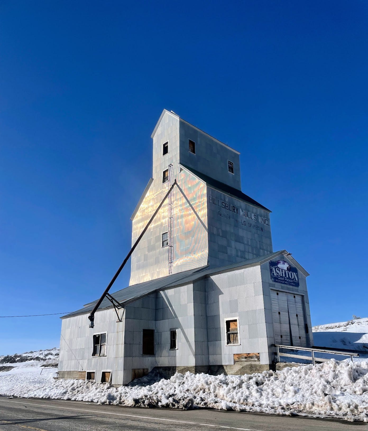 A metal grain silo reflects sunlight under a bright blue sky, surrounded by a field of snow.