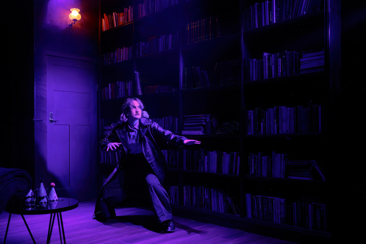 A photograph from the stage of the musical "Murder for Two". The character Detective Marcus strikes an action pose in front of a door and a large bookcase.