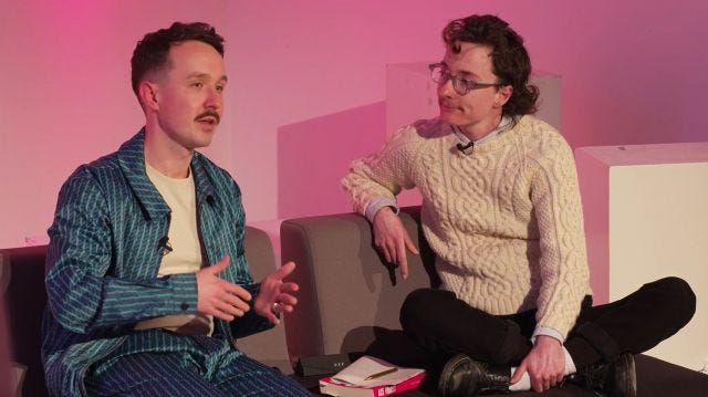 me listening to Kevin speak, we're both sitting on a sofa in front of a pink background. Kevin is wearing an incredible blue patterned satin tracksuit.