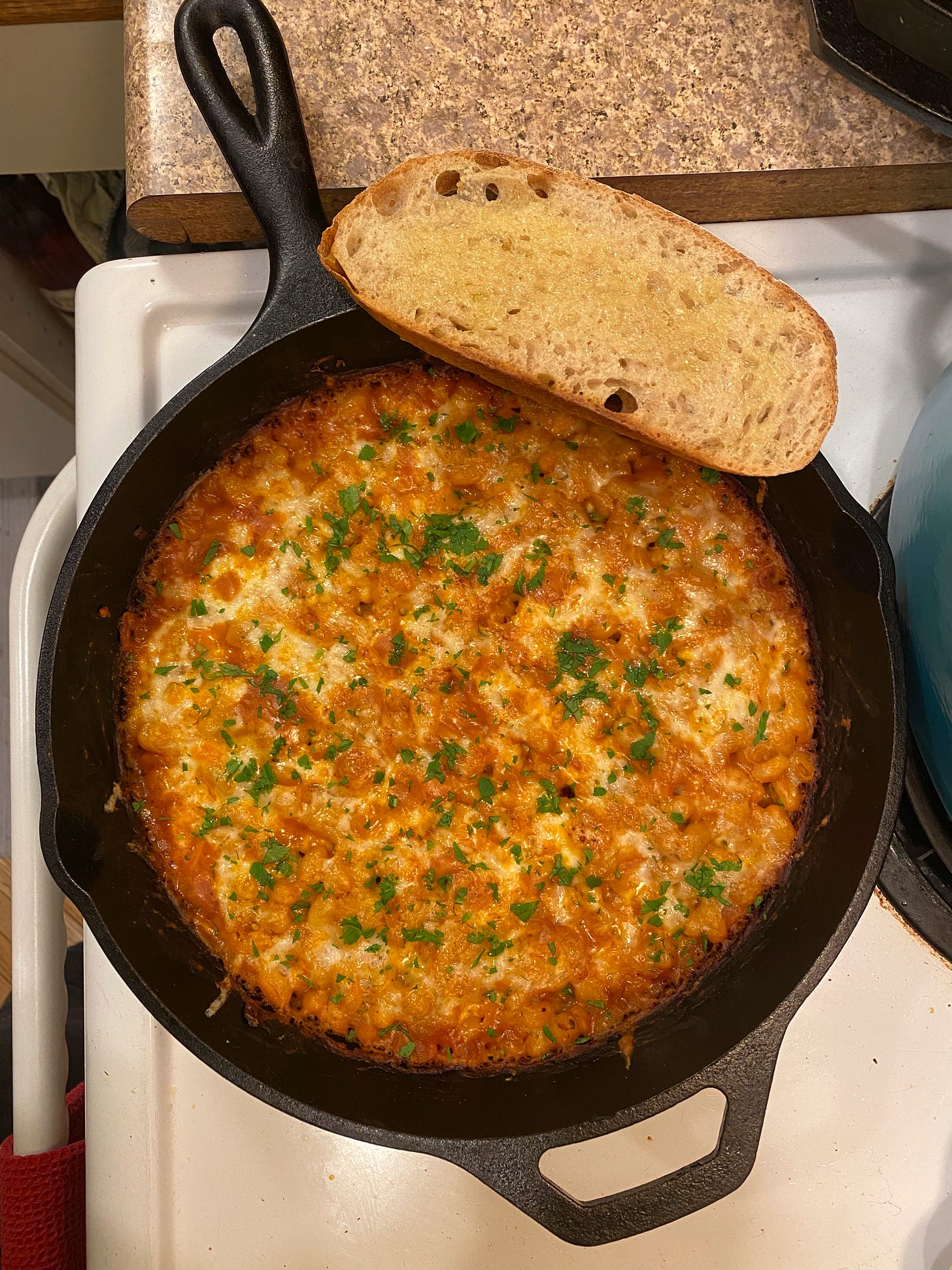 On the stove, from above, a cast iron pan full of cheesy baked beans. The tomato sauce has bubbled up over the cheese at the edges, and the dish is sprinkled with parsley. A slice of sourdough toast rests on the edge of the pan.
