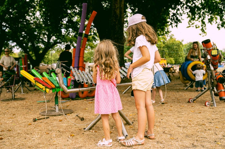 Two young girls playing on a large xylophone beneath a tree in a park