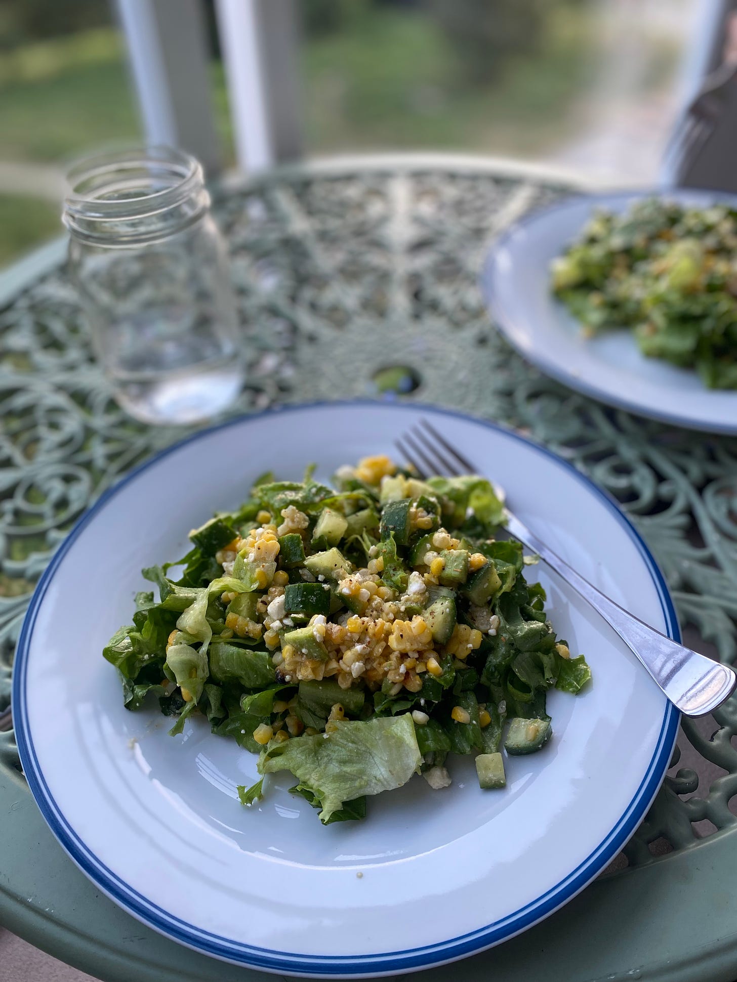 the corn salad described above, on a white plate with a blue rim and a fork resting at its edge. It's on a mint green café table next to a glass of water.