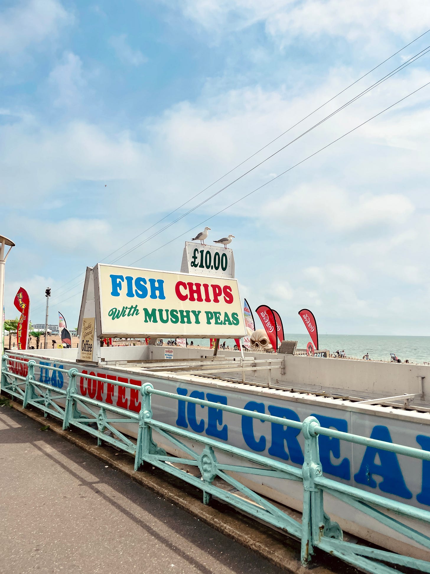 A colorful sign reads FISH CHIPS WITH MUSHY PEAS, on the beach in Brighton.