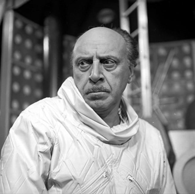 A late-middled-aged white man with a moustache and receding hairline. He wears a spacesuit without a helmet or breathing apparatus, and is within a space rocket.