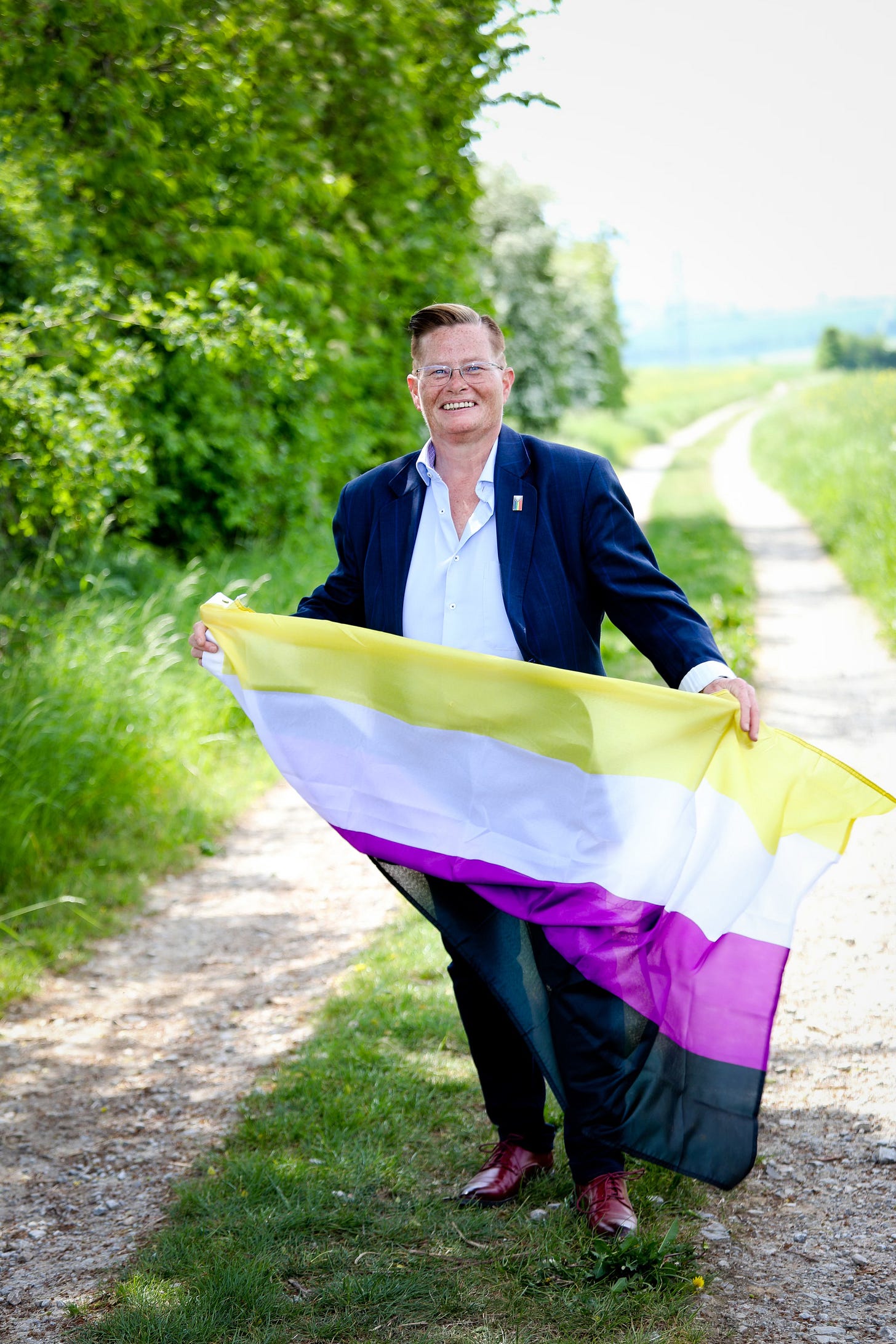  I am standing on a peaceful countryside path surrounded by greenery, smiling and holding the non-binary pride flag in yellow, white, purple, and black. I am dressed in a navy blazer, a white shirt, and red shoes, radiating confidence and happiness. Photocredit: Barbara Hötzel