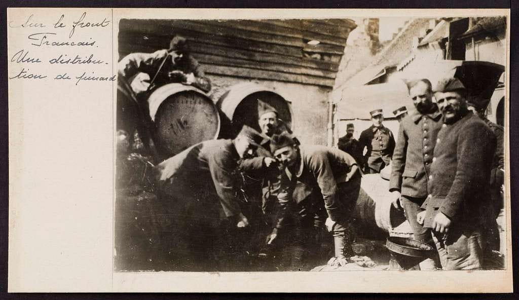 Some happy looking, sepia-toned, French soldiers are groups around a couple of barrels of wine. The caption reads, in French “Sur le front français. Une distribution de pinard. “