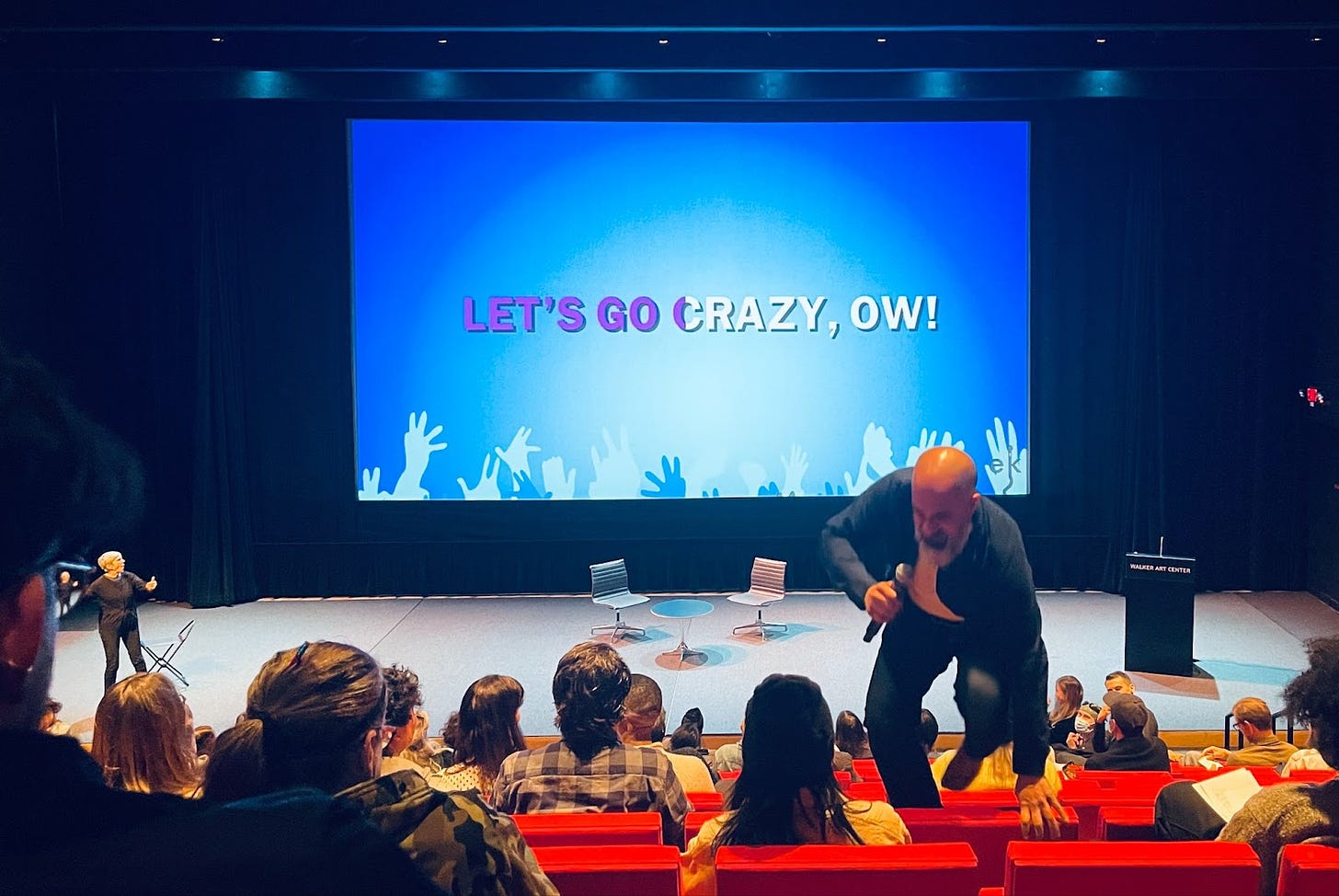 Large-scale auditorium with projection screen showing karaoke-style text, "LET'S GO CRAZY, OW!" Meanwhile, a bald, bearded man with a wireless microphone jumps over the audiences' seats as they look on, surprised.