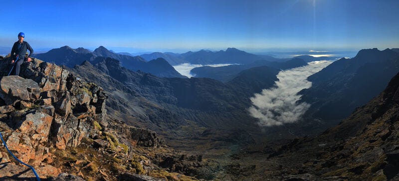 Panoramic view from the Cuillin on Skye. A man with a blue helmet is sitting on golden rocks overlooking the view. It is a beautiful day with clear, blue skies. The sun shines off the sea in the distance and there is some lens flair. The valley below is covered in fog but there is no cloud higher up.