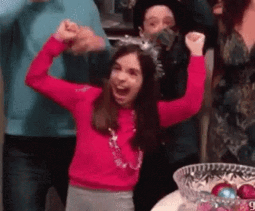 GIF of a young brunette girl in a pink long sleeve shirt and party hat slamming a glass bowl onto the ground before screaming and lifting her arms into the air in triumph. she is surrounded by screaming white people