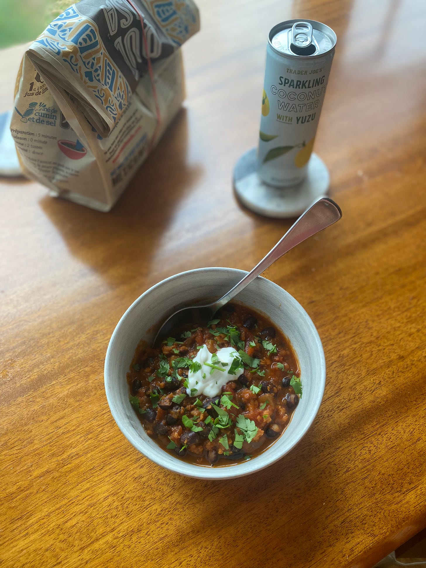 a bowl of the chili described above, topped with cilantro and sour cream. In the background is a bag of tortilla chips with a rubber band around it, and a can of sparkling coconut water on a coaster.