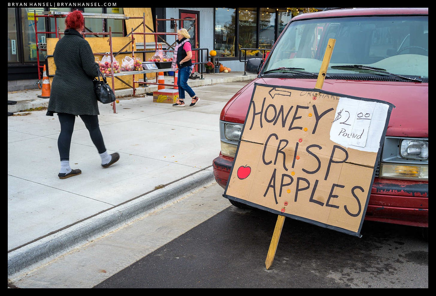 A sign that says, "Honey Crisp Apples" leaning against a red van. On construction scaffolding bags of apples sit. Two people walk past. One woman's hair is the same red as the van. 