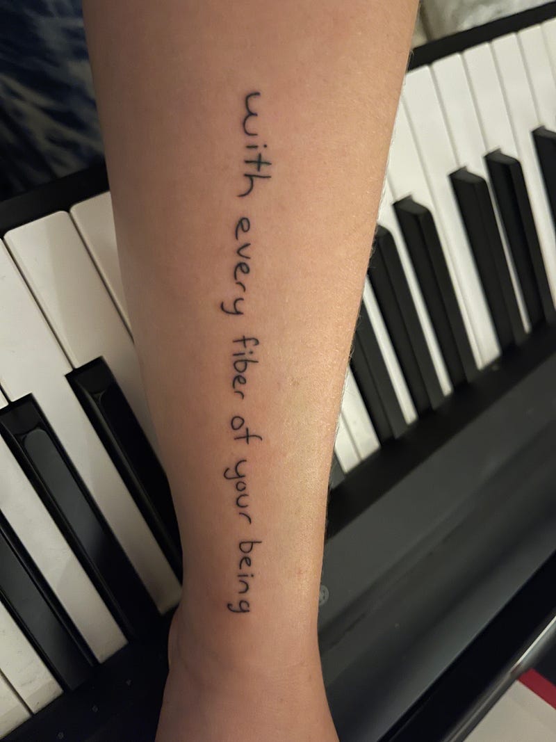 forearm placed over piano keys bearing a tattoo in printed handwriting with text “with every fiber of your being”