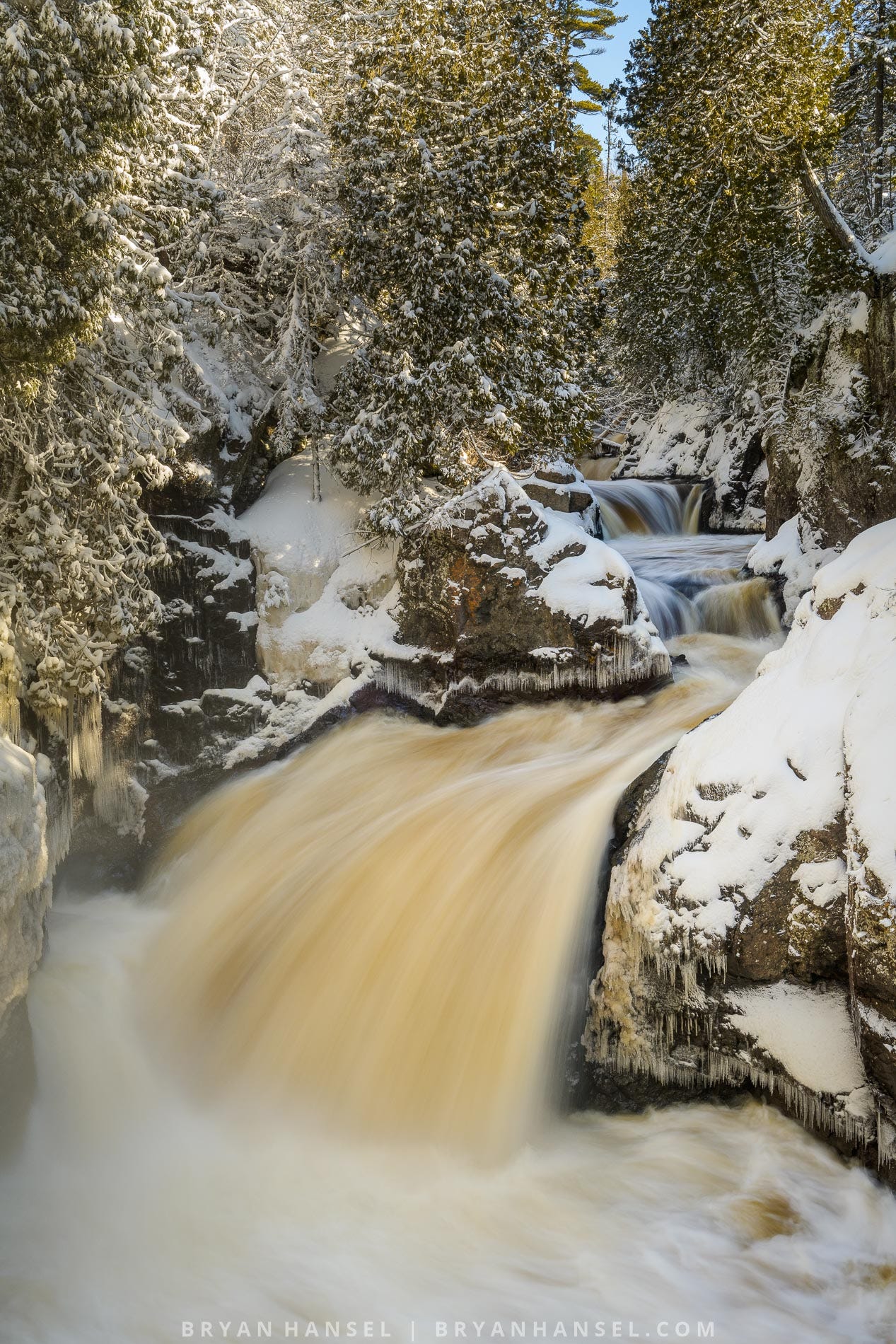 A waterfall surrounded by snow-covered trees and rocks. There's blue sky in the background.