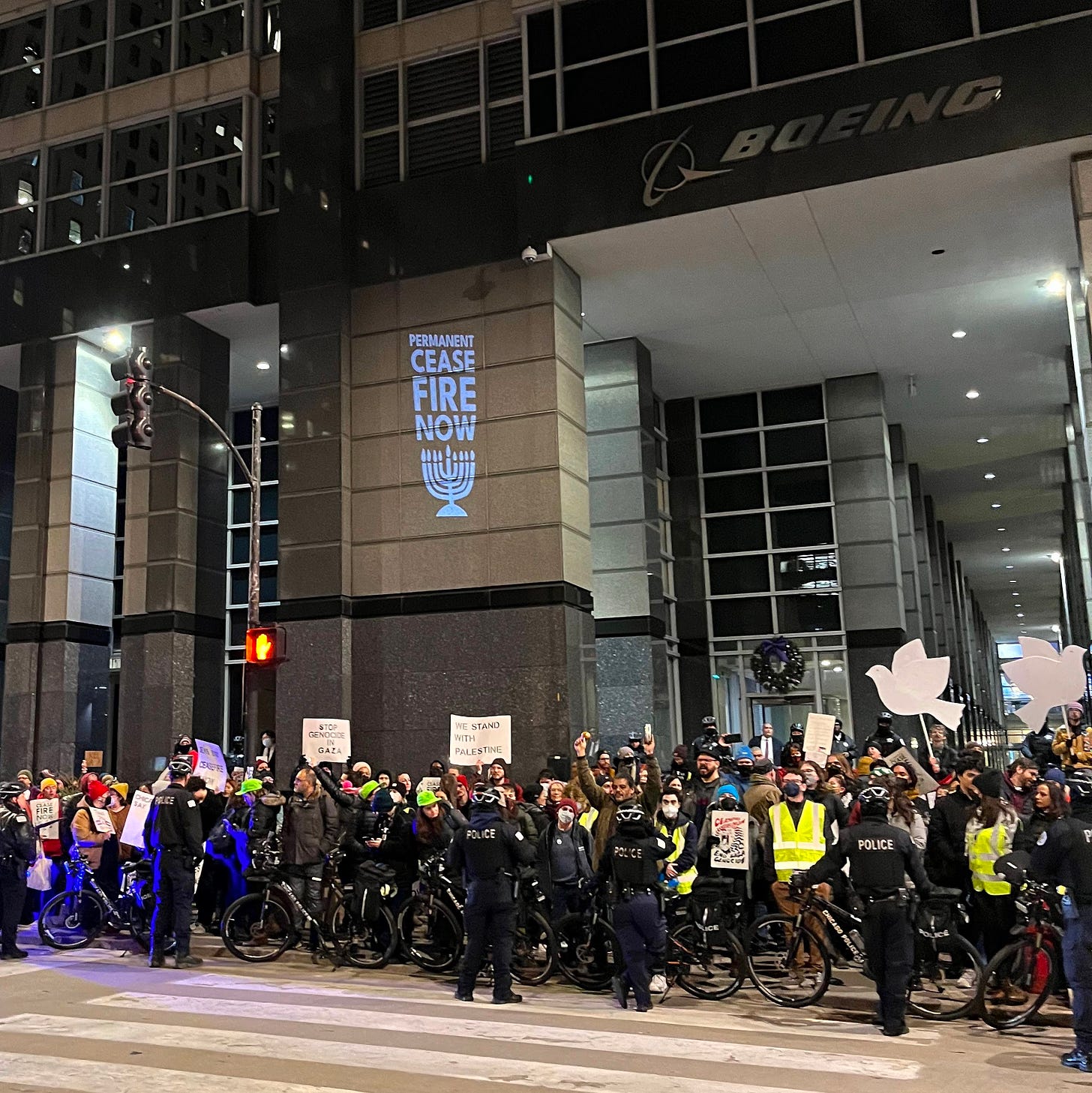 A message that reads "Permanent Ceasefire Now" above an image of a menorah is projected on the Boeing building in Chicago. There are hundreds of protesters in Chicago and a line of police blocking them from entering the street.