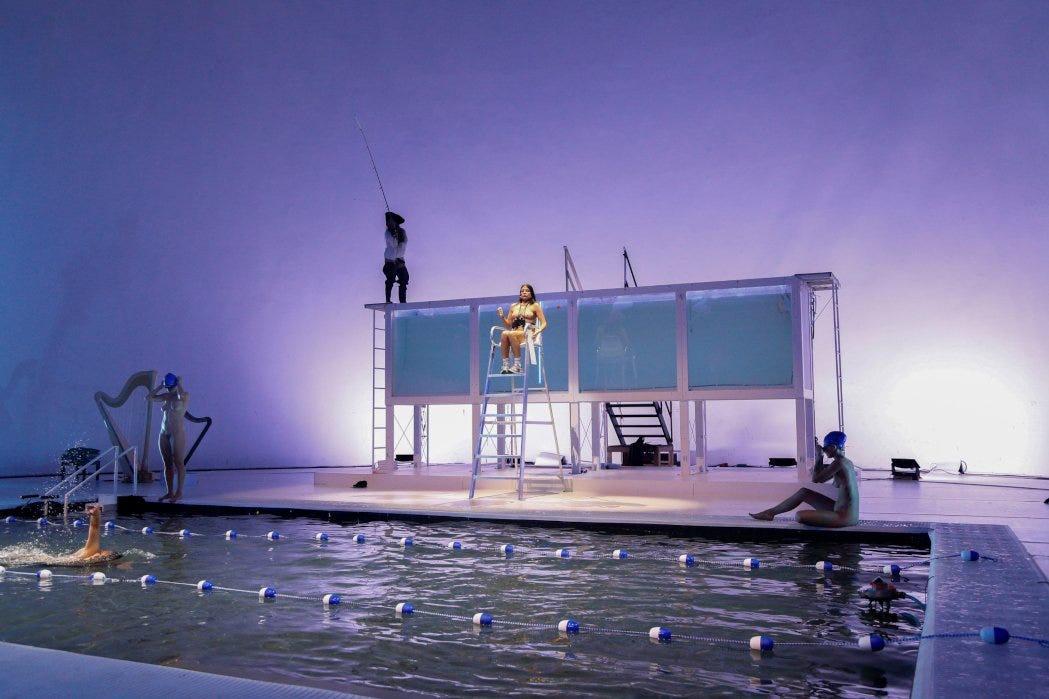 Imagem: Palco com uma piscina esportiva no meio, uma mulher nadando, uma sentada ao lado, outra em pé se preparando para pular. Uma escada de bombeiro com uma mulher sentada no alto. Atrás há uma caixa retangular como um aquário com agua azul clara. Nela, há uma mulher em pé pescando.