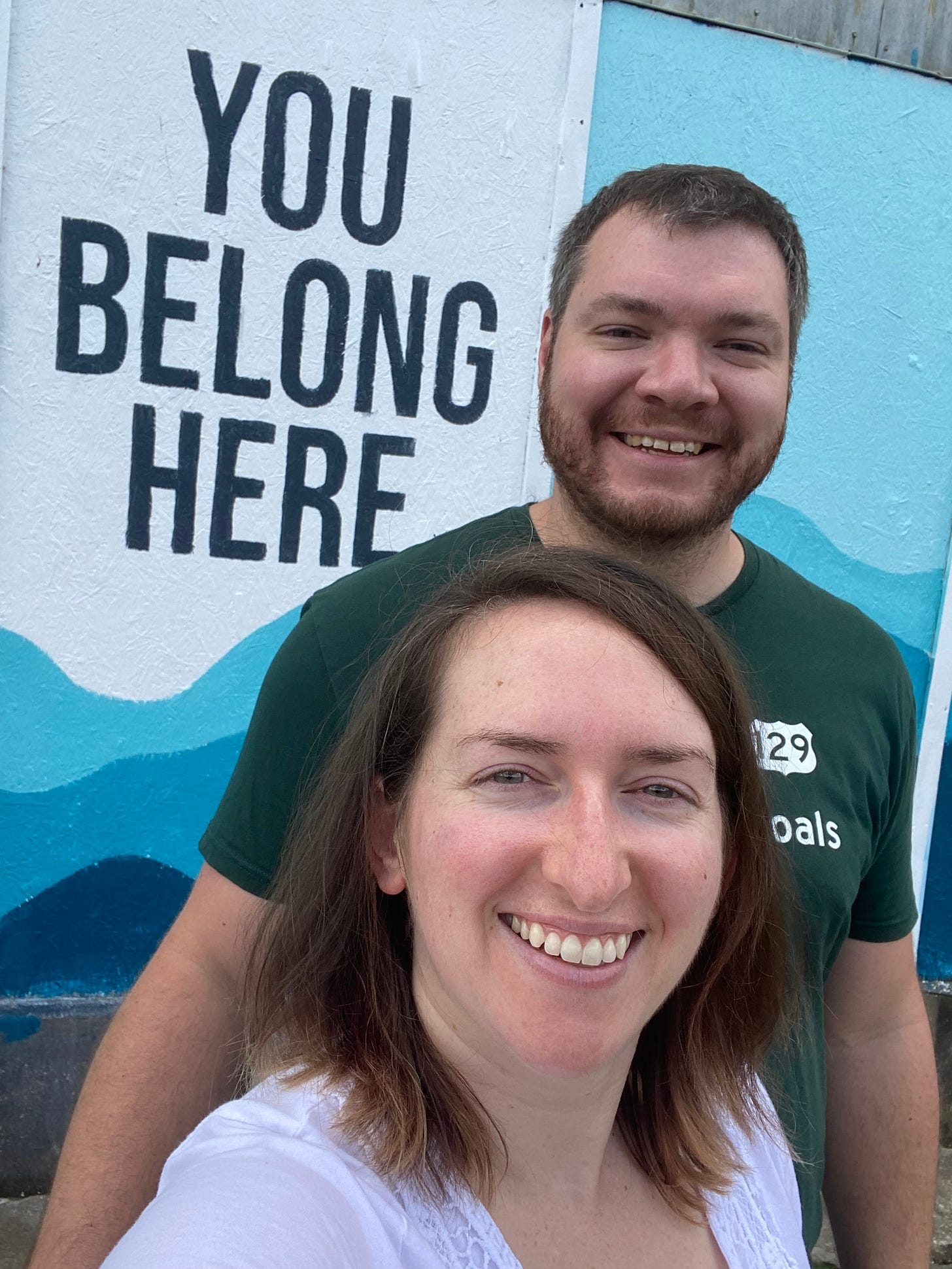 smiling white man and smiling white woman in front of a mural that says "You Belong Here"