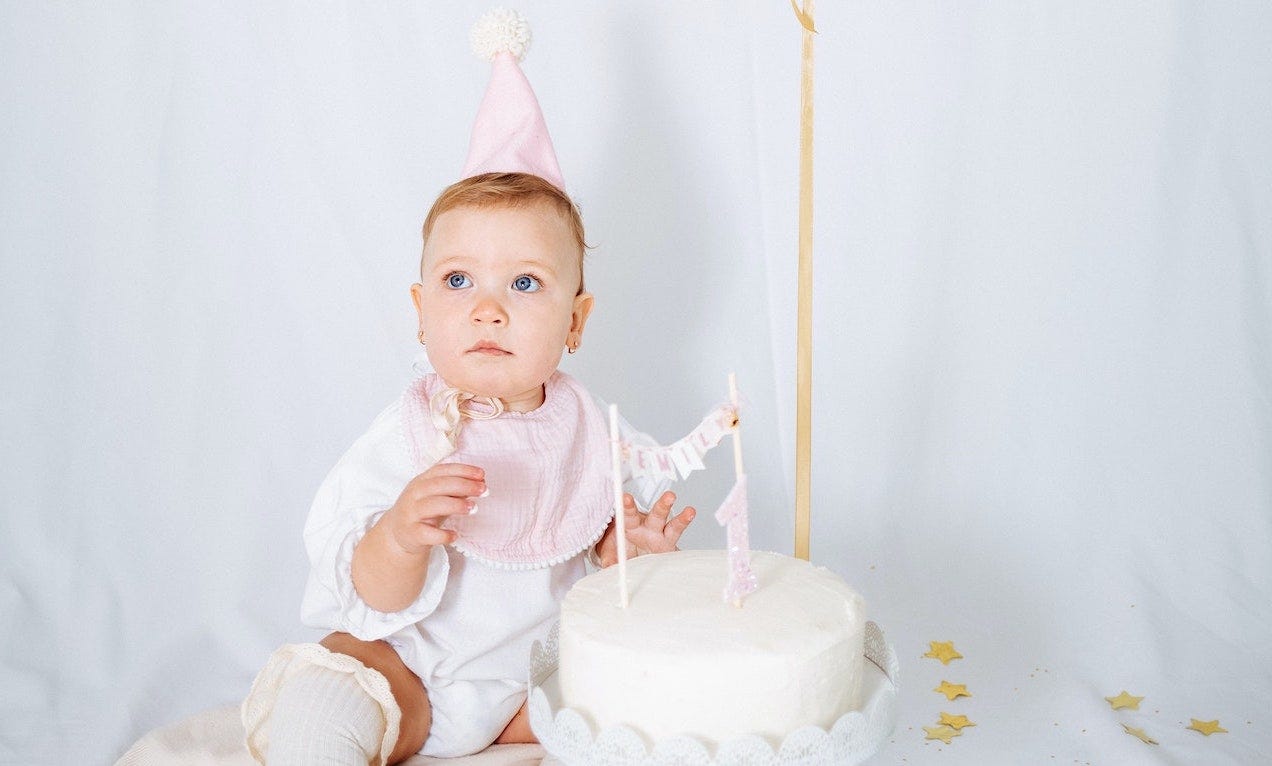 Baby in birthday hat sitting by a white cake