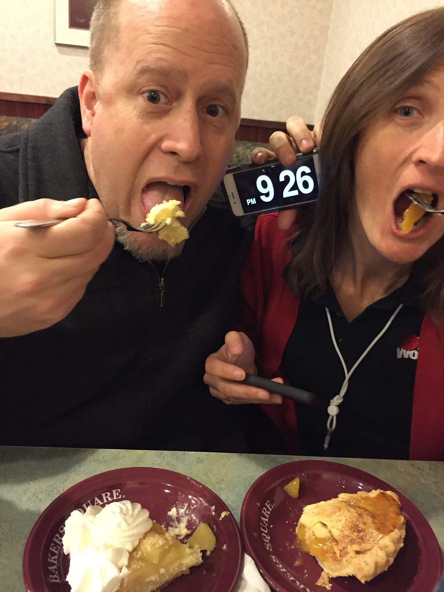 Andy and I shoveling pie into our faces at a restaurant while holding up a phone that shows the time to be 9:26.