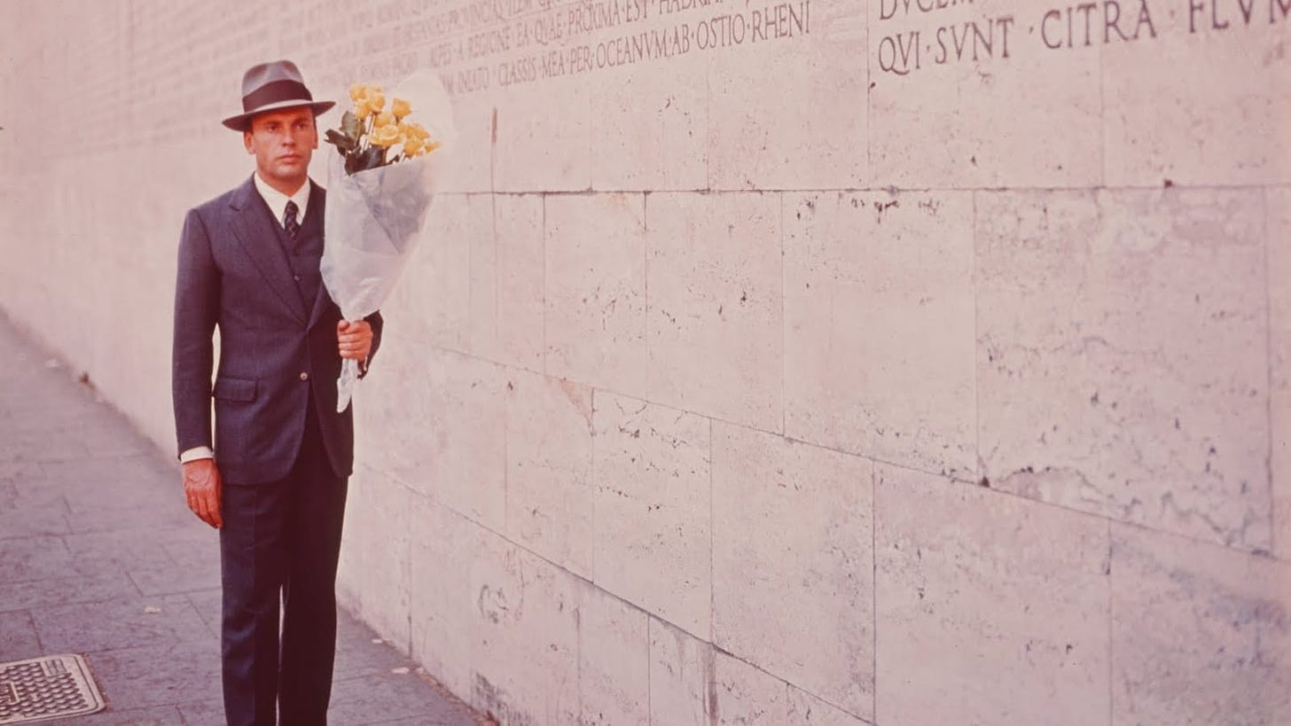 The Conformist | Still features Jean-Louis Trintingnant as Marcello Clerici carrying a bouquet against a white wall.