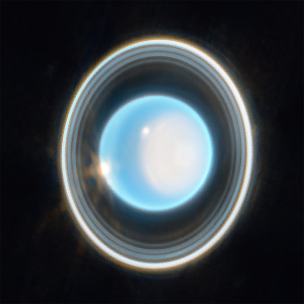 The planet Uranus vertical rings on a black background. The planet is light blue with a large, white patch on the right side. On the edge at the upper left is a bright white spot, with another white spot on the left side of the planet.