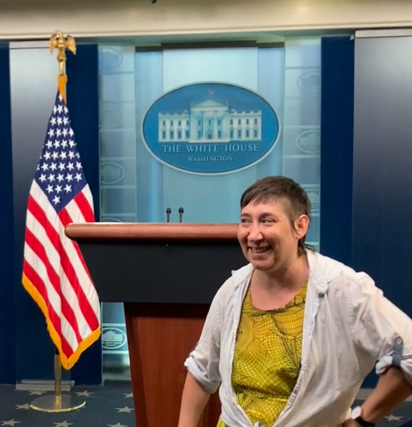 me, a white person, wearing a yellow dress and a blue button up shirt, in front of the white house press pool room