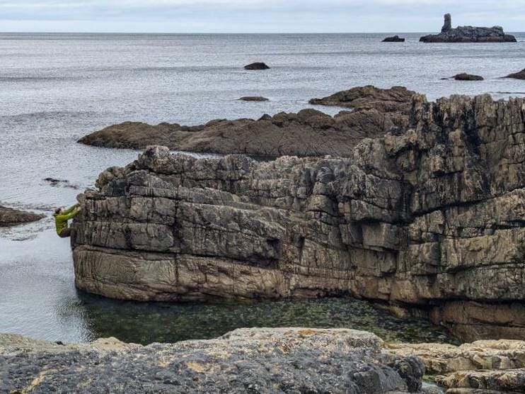 The view shows a rocky coastline with 5 m high cliffs behind an inlet of sea. There is more sea in the background and some sea stacks. On the left-most end of the band of cliffs in the mid-distance is a man in a yellow jacket climbing on the rocks above the sea. His figure is outlined by the sea behind. How did he get there?