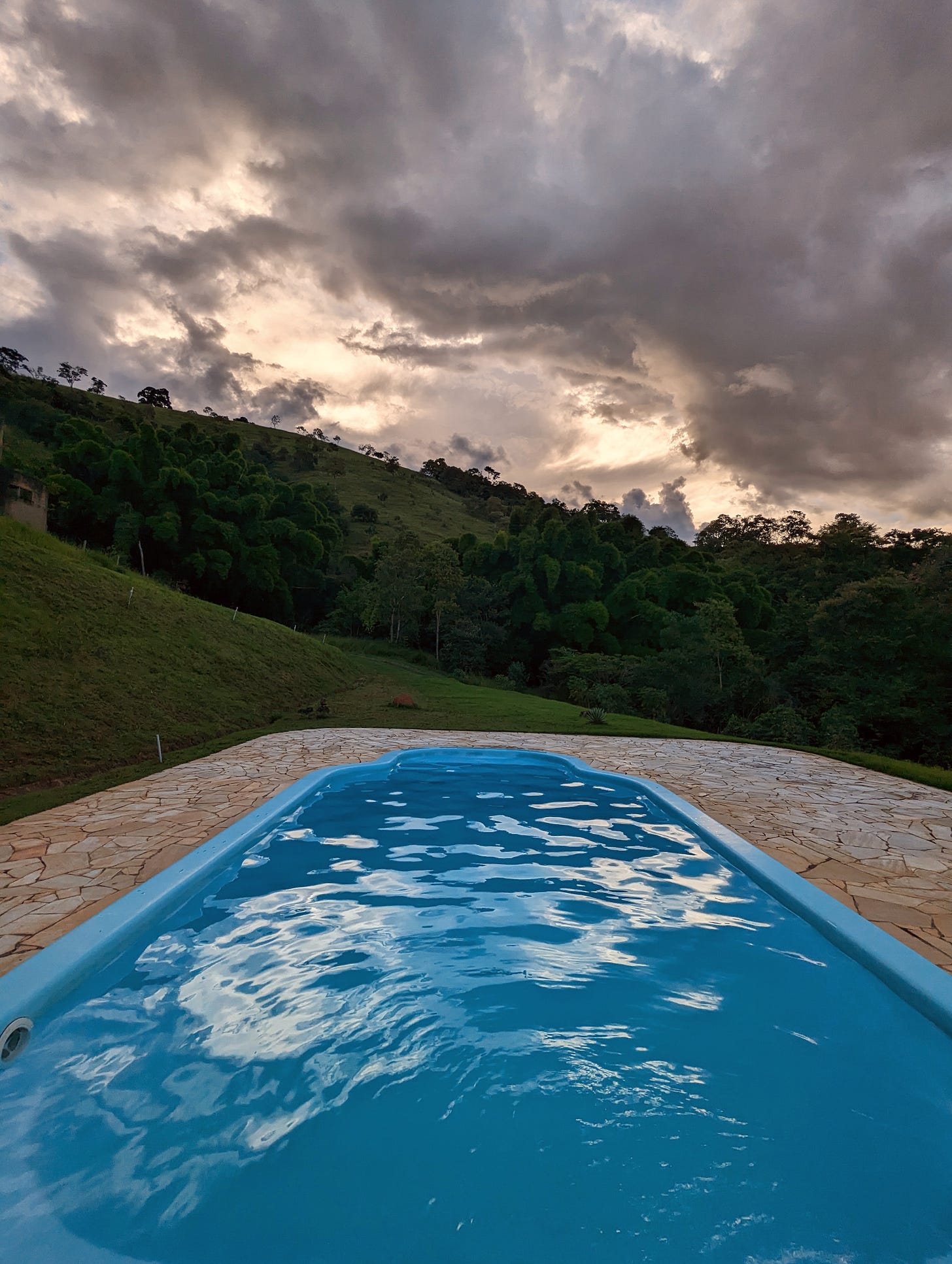 A swimming pool surrounded by steep hills with some trees, a sky heavy with dark clouds above it.