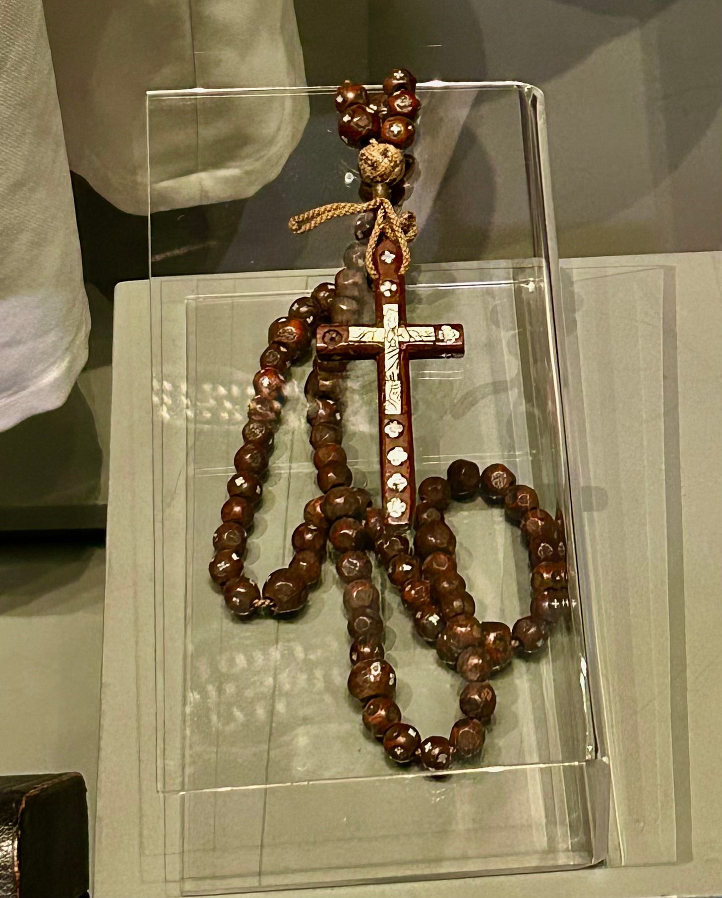 A wooden rosary, decorated with silver religious symbols. The beads are also wooden with some decoration which is not entirely clear due to the glass reflections - the rosary is held within a glass case.