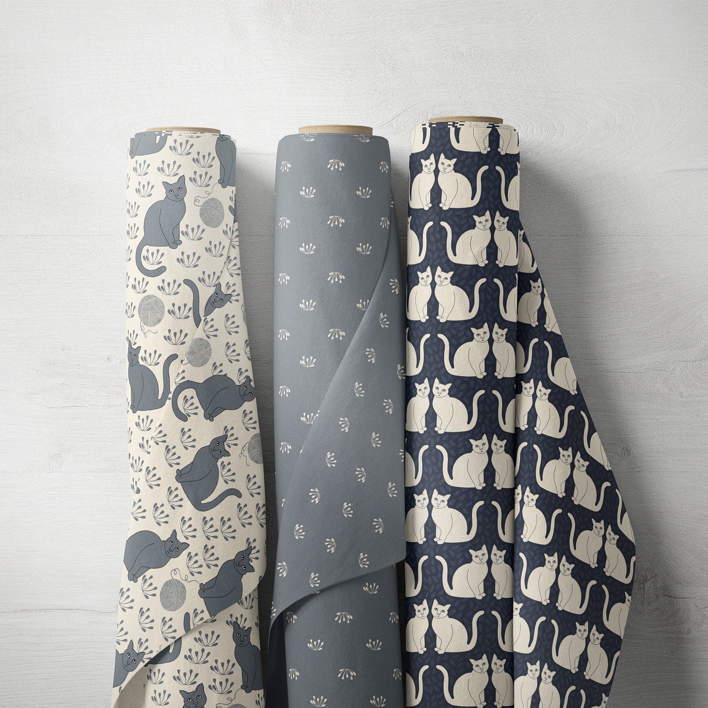 Photograph of three fabric rolls leaning upright against the wall. The left fabric roll depicts a pattern of slate grey cats on a white background, playing with yarn in a field of grasses. The center fabric roll is a coordinate pattern, slate grey with  a half-drop botanical pattern. The right fabric roll is a repeat pattern of two seated white cats facing each other, on a dark blue background surrounded by slightly lighter blue paintstrokes.