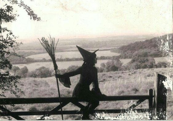 This may contain: an old photo of a woman dressed as a witch holding a broom and standing on a fence