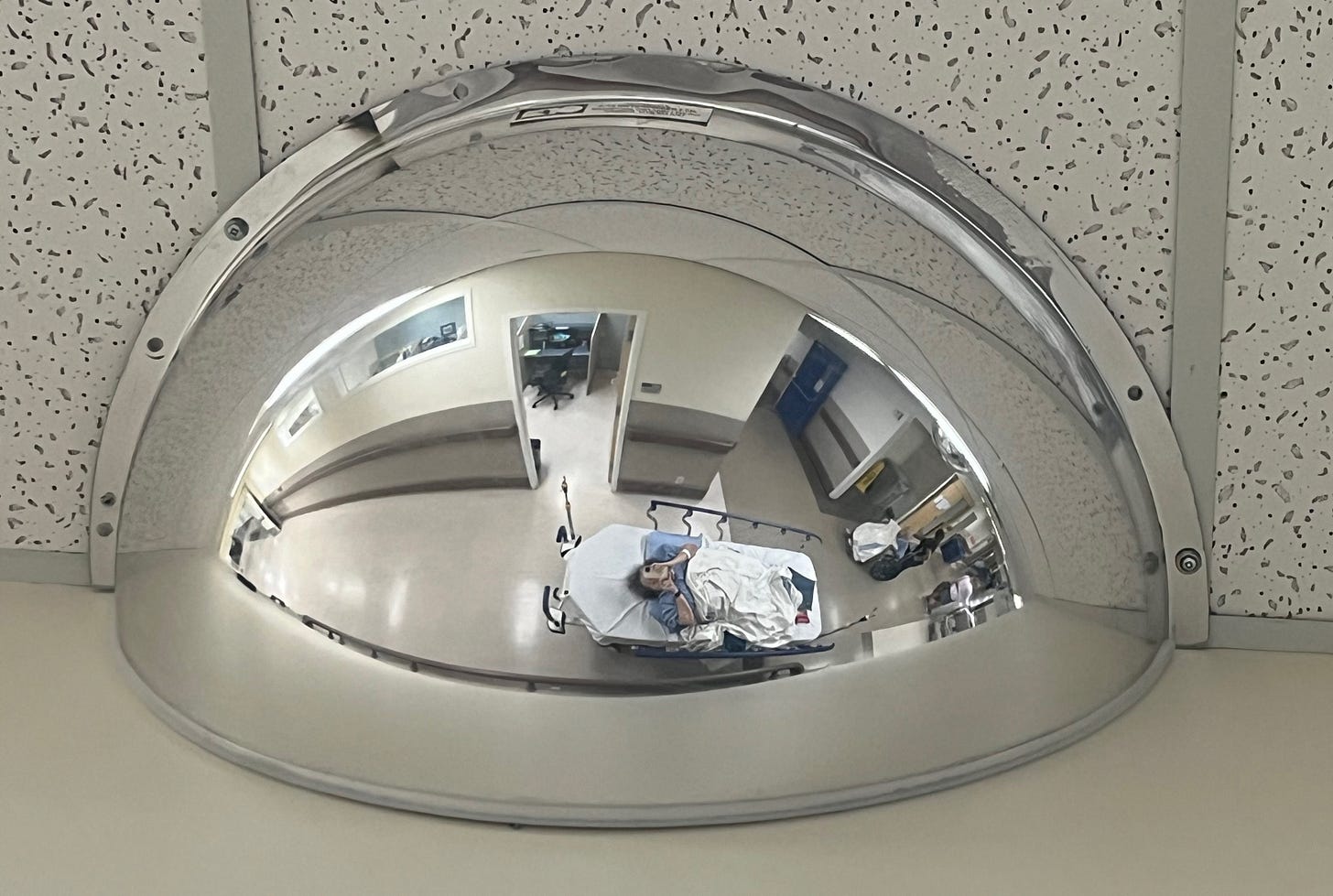 Photo of a half sphere mirror on the ceiling in the hallway of a hospital. Athena appears in the reflection taking the photo from her back on the stretcher.